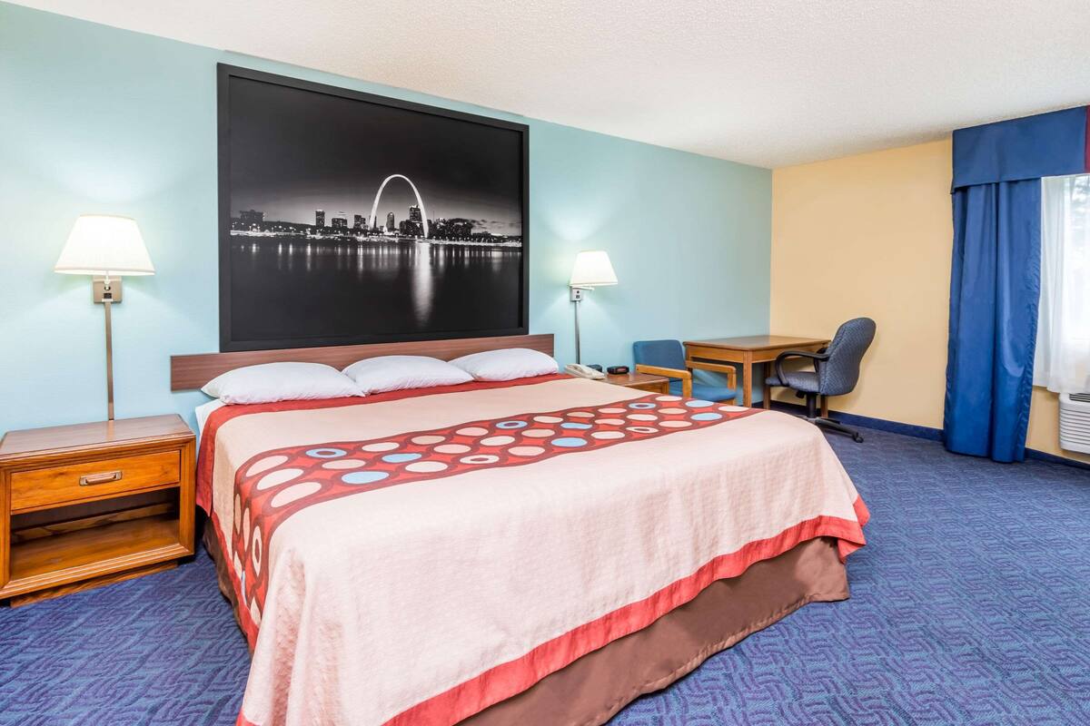 A hotel room with a bed, two bedside tables, a desk, a chair, and a large cityscape painting on the wall.