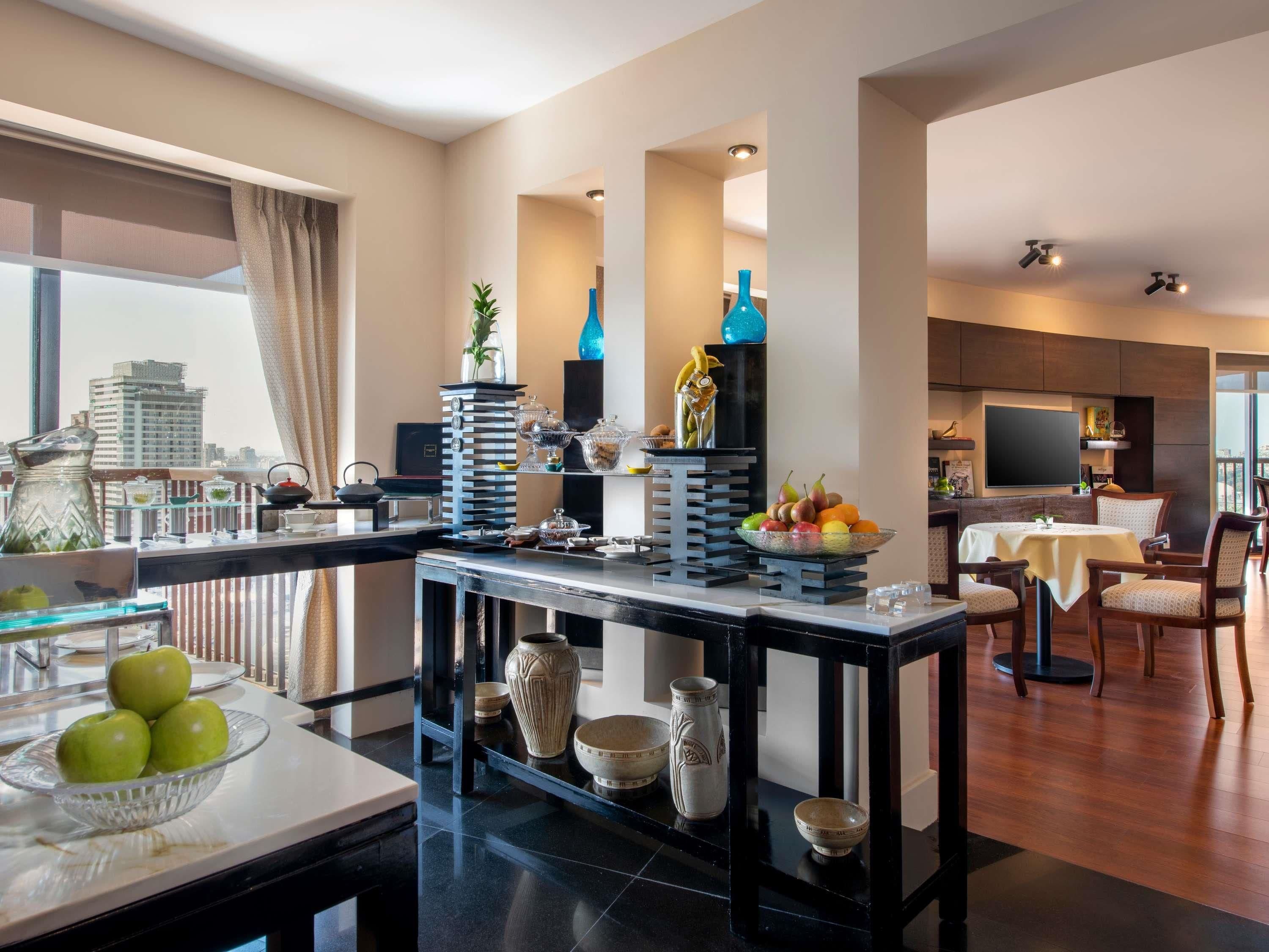 A modern kitchen with a dining area, featuring a large window, a fruit bowl, and a variety of kitchenware.