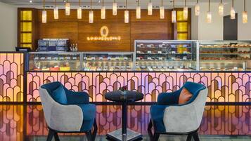 Un café avec un comptoir en bois où sont exposées des pâtisseries et une machine à café, deux chaises bleues et des lampes suspendues.