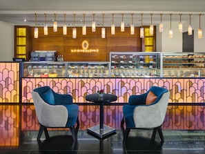 A café with a wooden counter displaying pastries and a coffee machine, two blue chairs, and hanging pendant lights. - Sofitel Cairo Nile El Gezirah (Cairo)