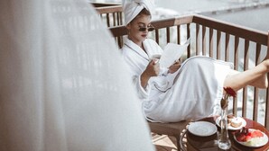 A person in a white bathrobe sitting on a wooden deck, reading a book. There is a plate with food and a glass of water on the table.