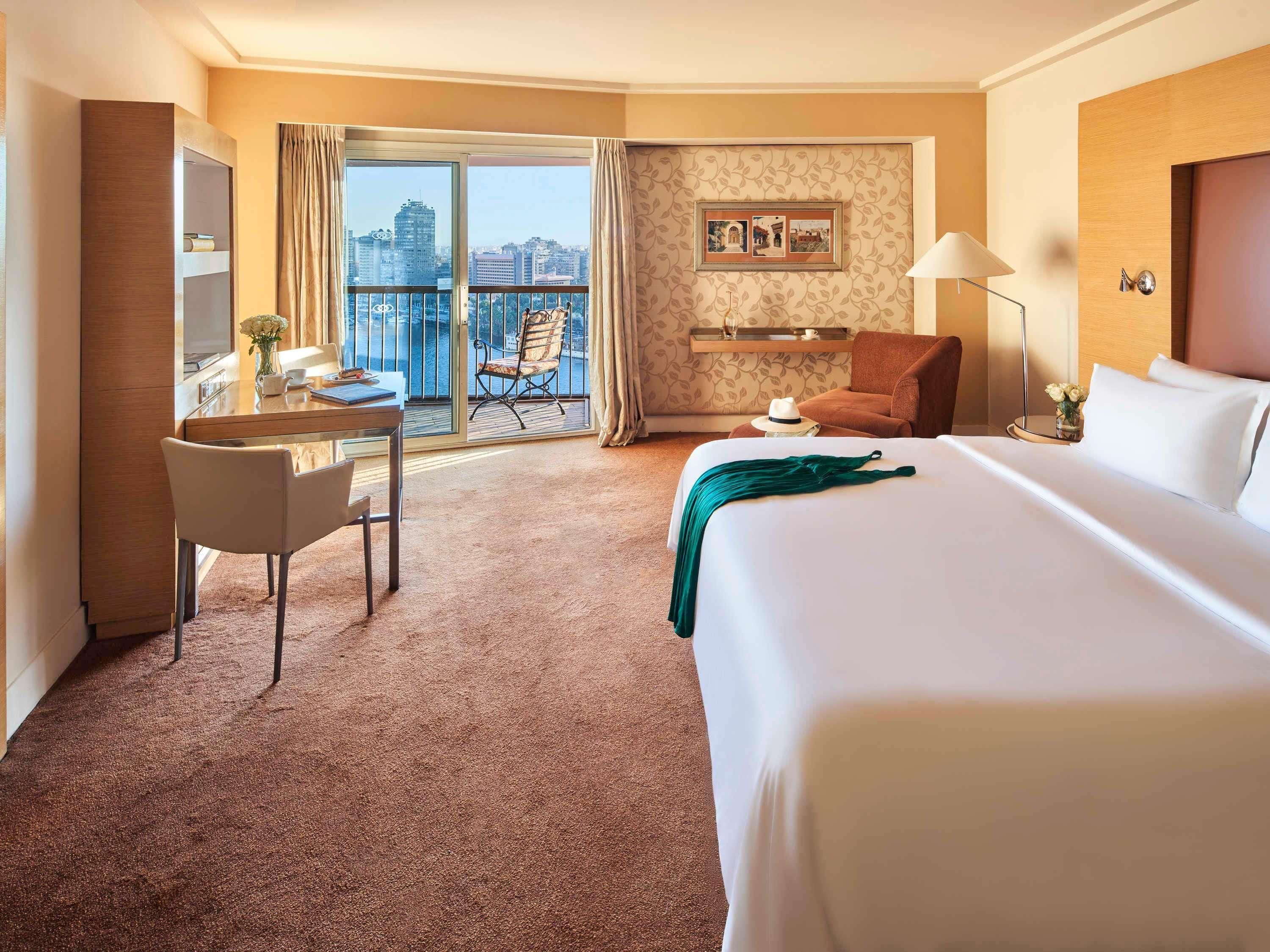 A hotel room with a bed, a desk, a chair, a balcony with a view of the city, and a wall with a framed picture.