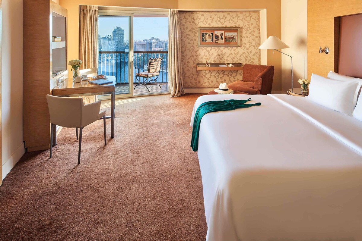A hotel room with a bed, a desk, a chair, a balcony with a view of the city, and a wall with a framed picture.