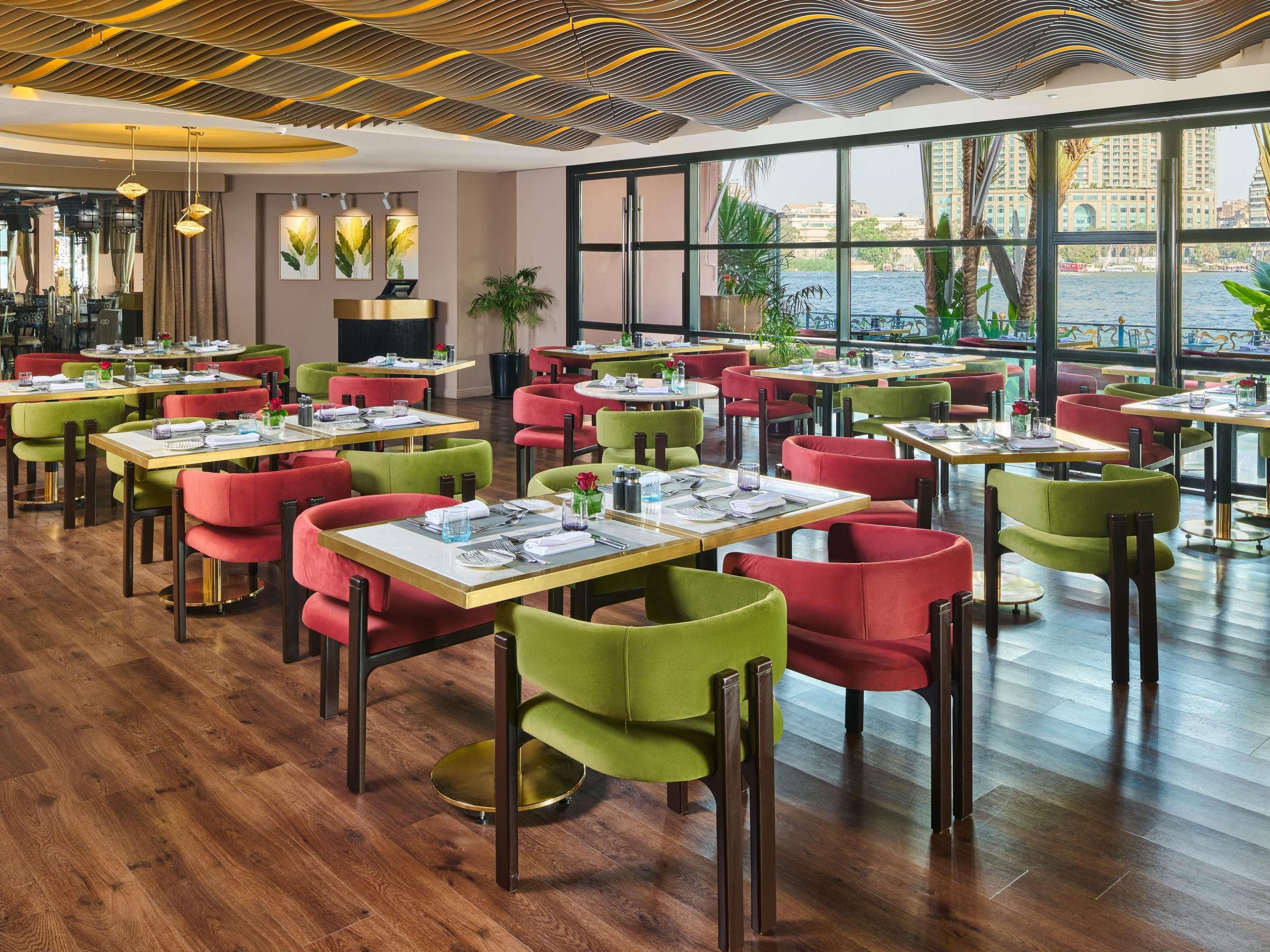 Un ristorante con diversi tavoli e sedie, ampie finestre che offrono vista su edifici e sull'acqua, e un design moderno del soffitto.