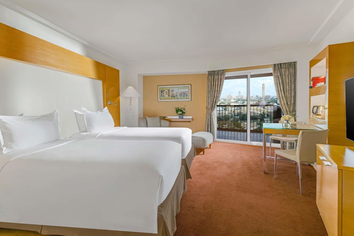 A hotel room with two beds, a desk, a chair, a TV, a balcony with a city view, and a small table with a lamp.