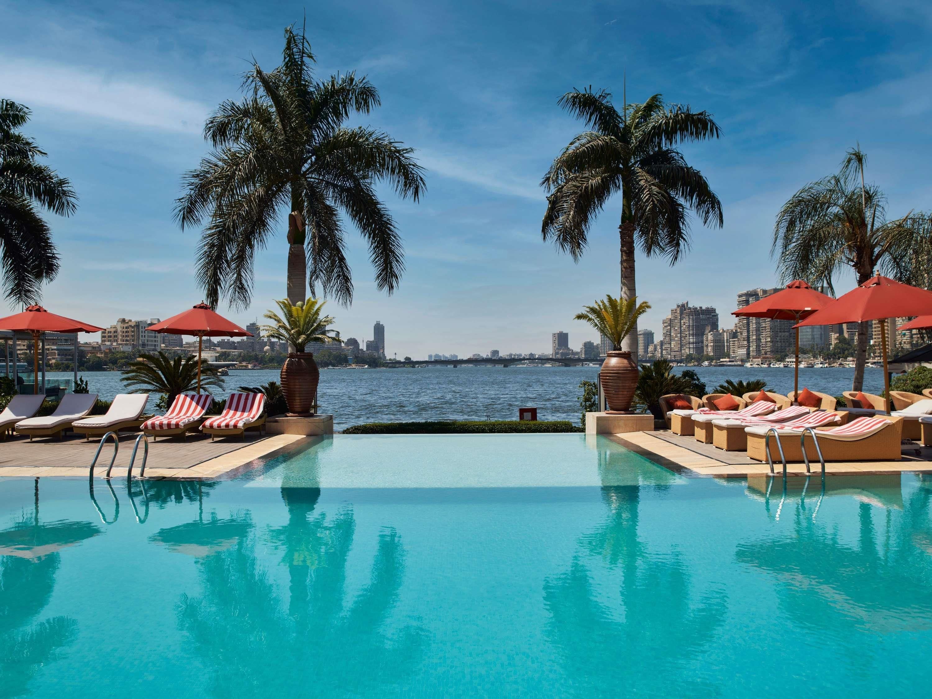 A pool area with lounge chairs and umbrellas, palm trees, and a view of a city skyline across a body of water.