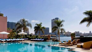 A pool area with lounge chairs and umbrellas, a clear view of the city skyline, and a calm water surface. - Sofitel Cairo Nile El Gezirah (Cairo)