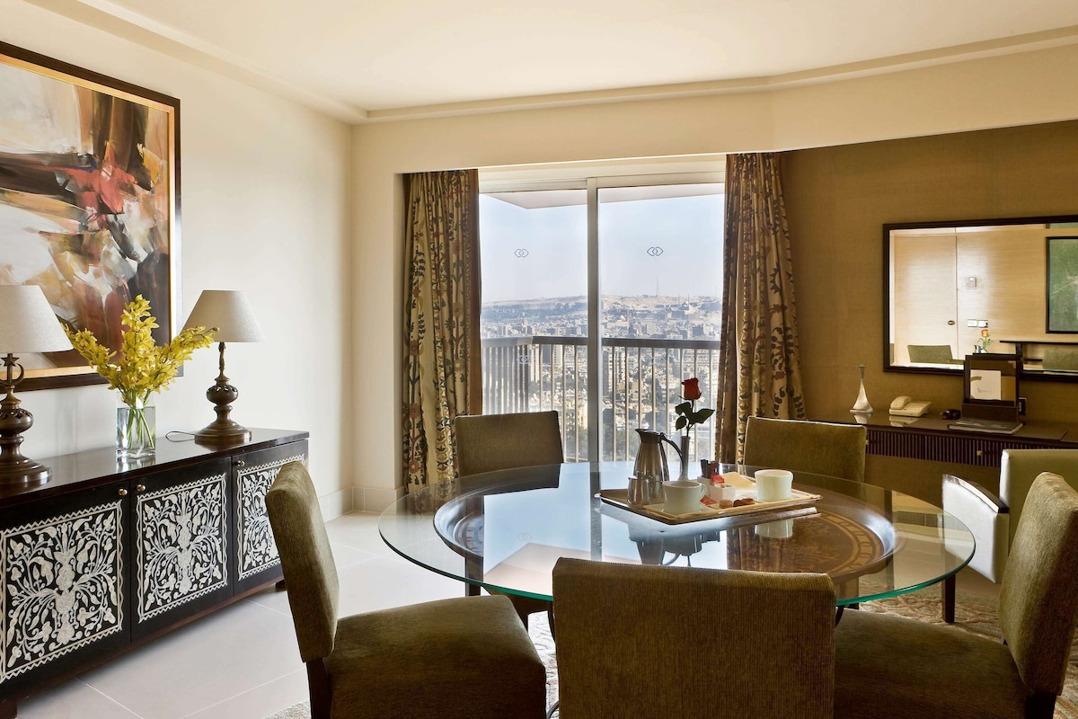 A modern hotel room with a glass dining table, comfortable seating, a decorative vase, and a city view through large windows.