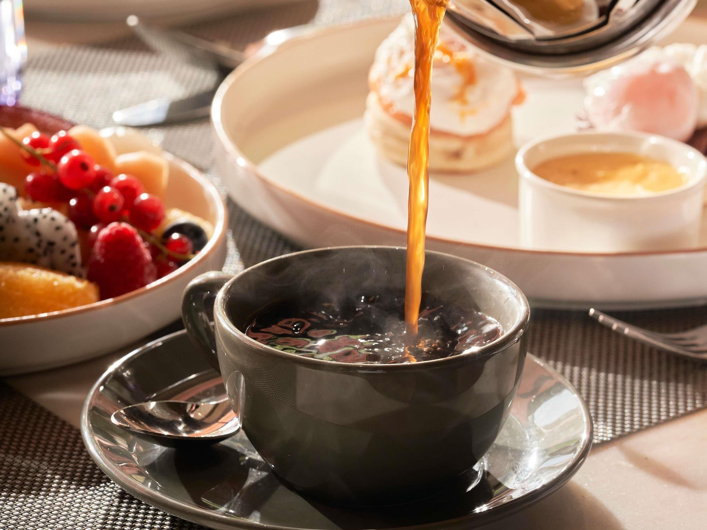 A cup of hot beverage on a saucer, a plate with pancakes, and a bowl of fruit.