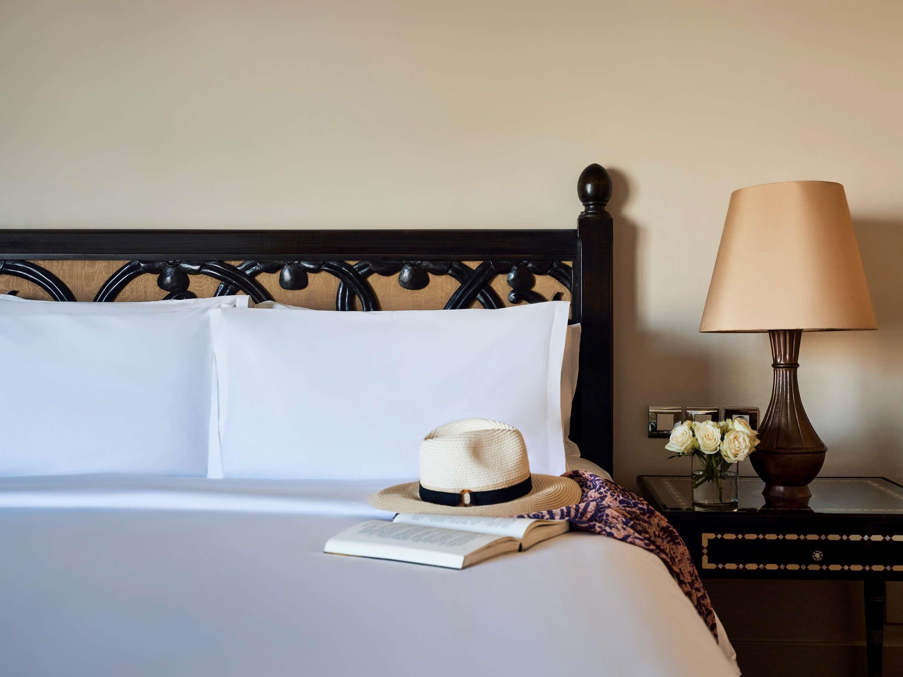 A neatly made bed with white linens, a book, a straw hat, and a lamp on a bedside table with a vase of flowers.
