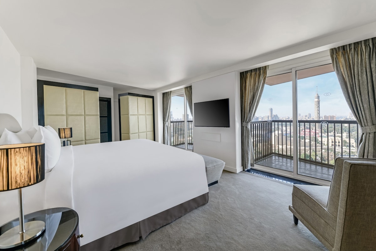 A hotel room with a large bed, a TV mounted on the wall, a balcony with a city view, and a sofa.