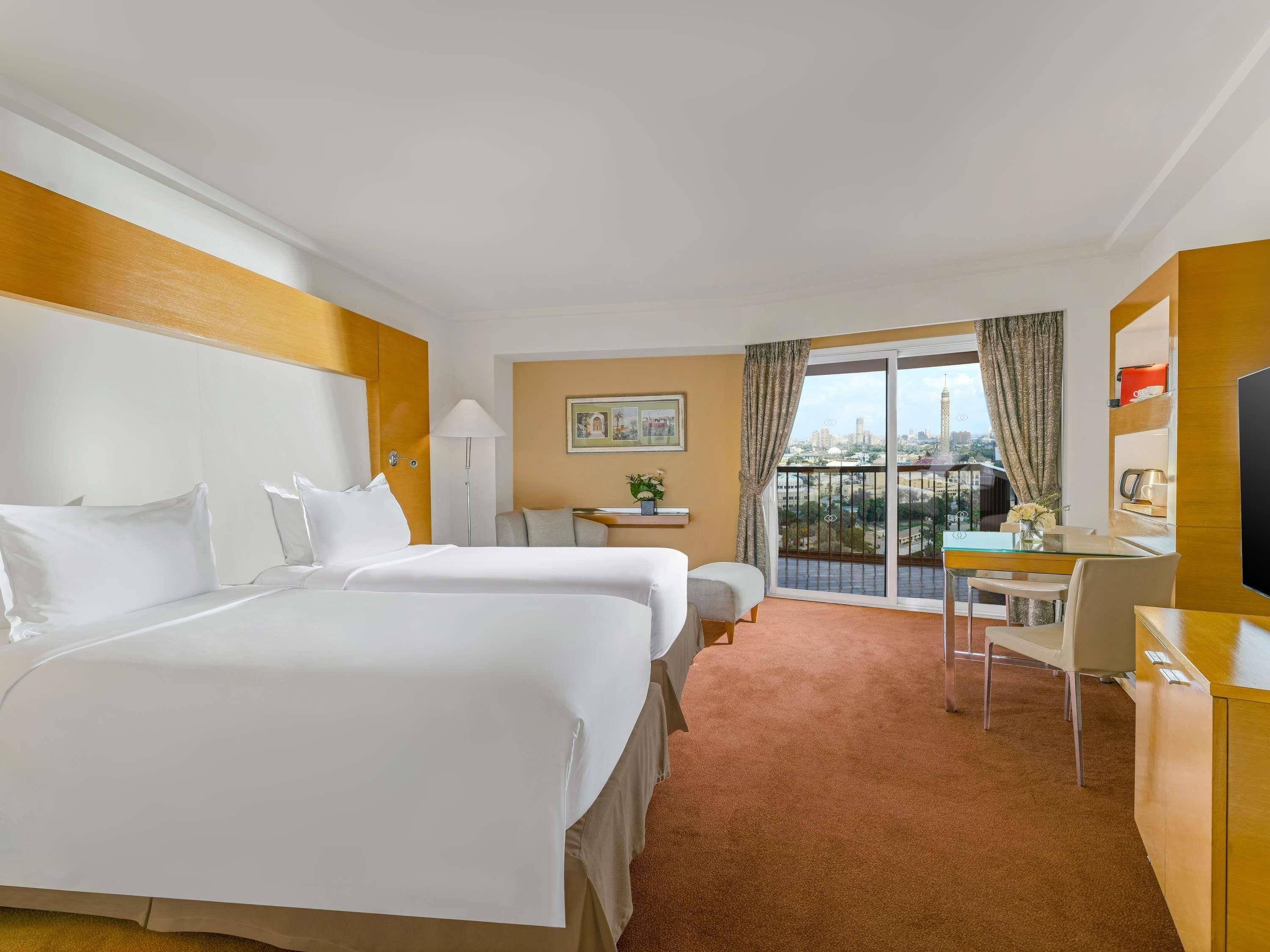 A hotel room with two beds, a desk, a chair, a TV, a balcony with a city view, and a small table with a lamp.