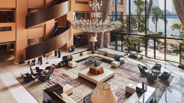 A spacious lobby with a grand staircase, a chandelier, and a large window offering a view of palm trees and a body of water.