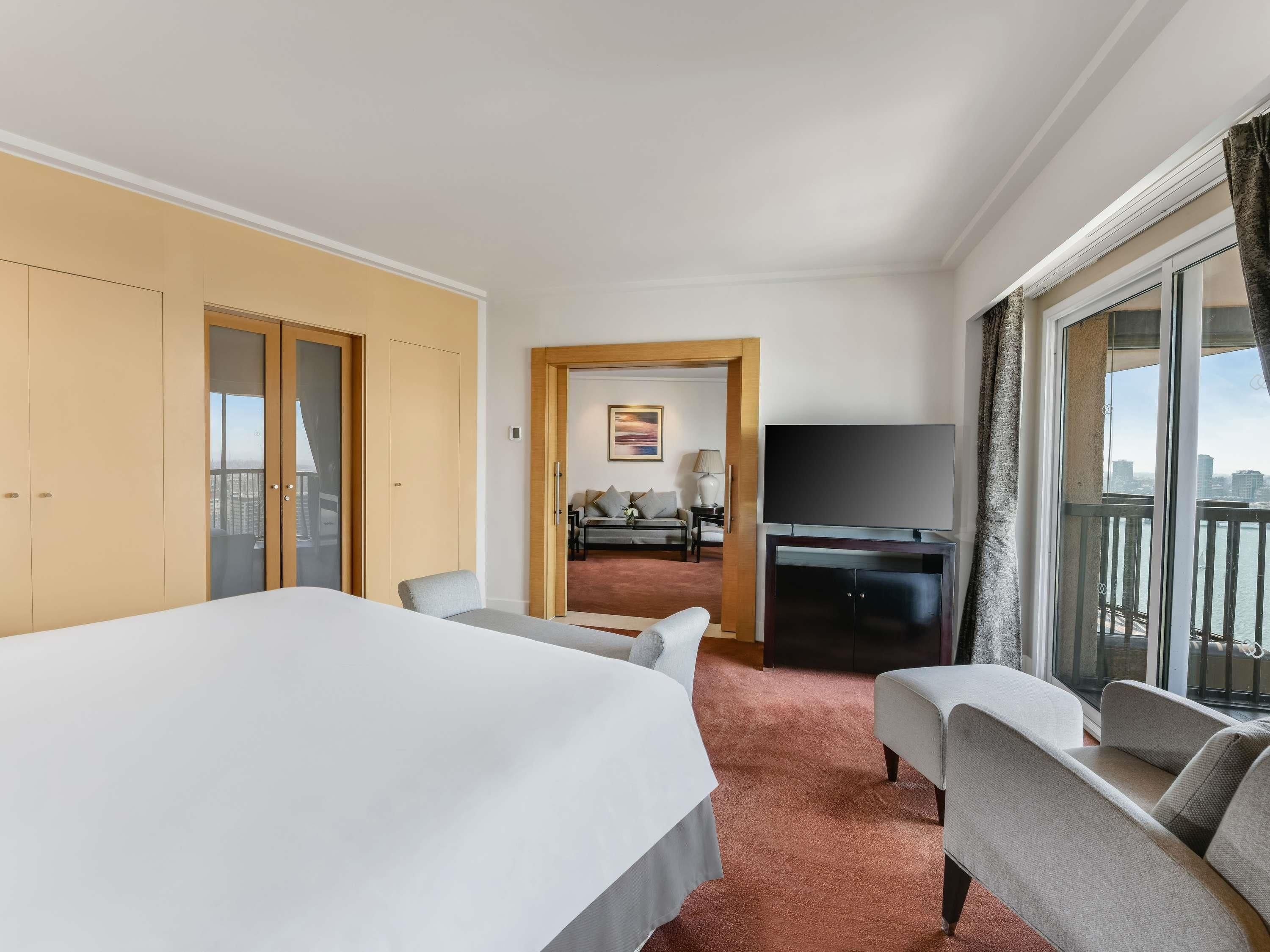 A hotel room with a large bed, a TV, a sofa, and a balcony with a city view.