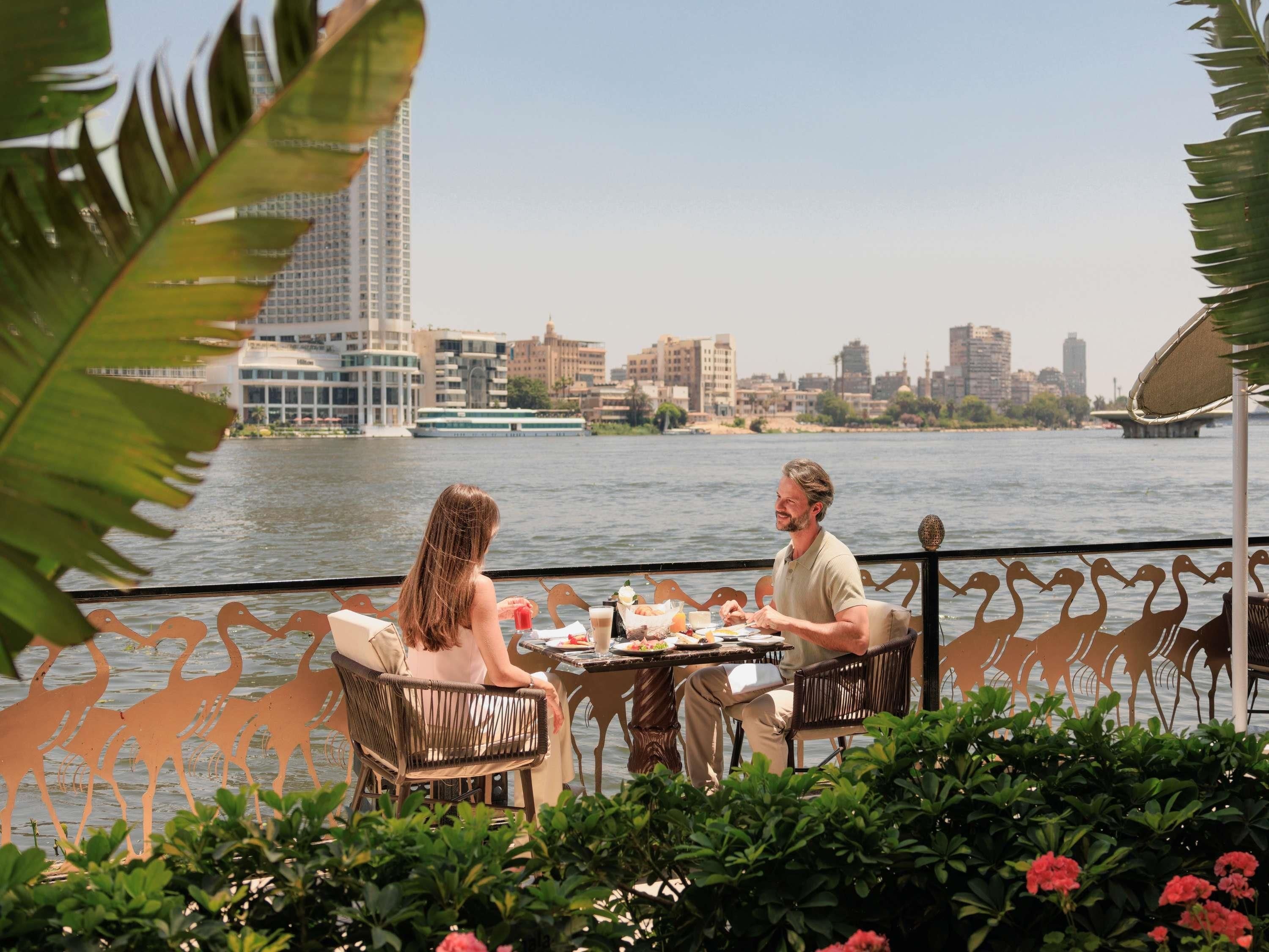 A dining table with chairs, a view of a river, and buildings in the background.