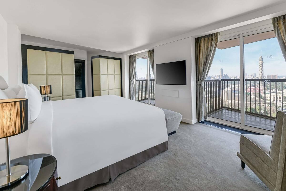 Habitación de hotel con una cama grande, televisión, balcón con vistas a la ciudad y ventana con cortinas.