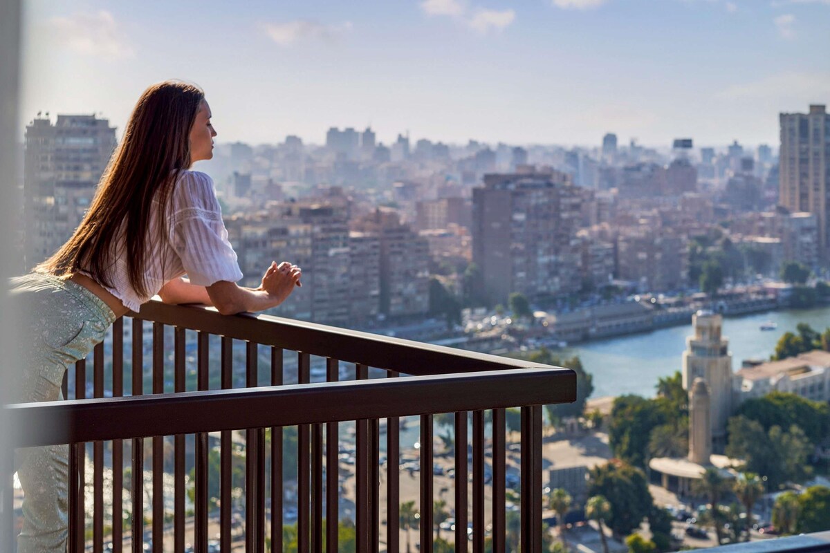A person sitting on a balcony overlooking a cityscape with buildings and a river.