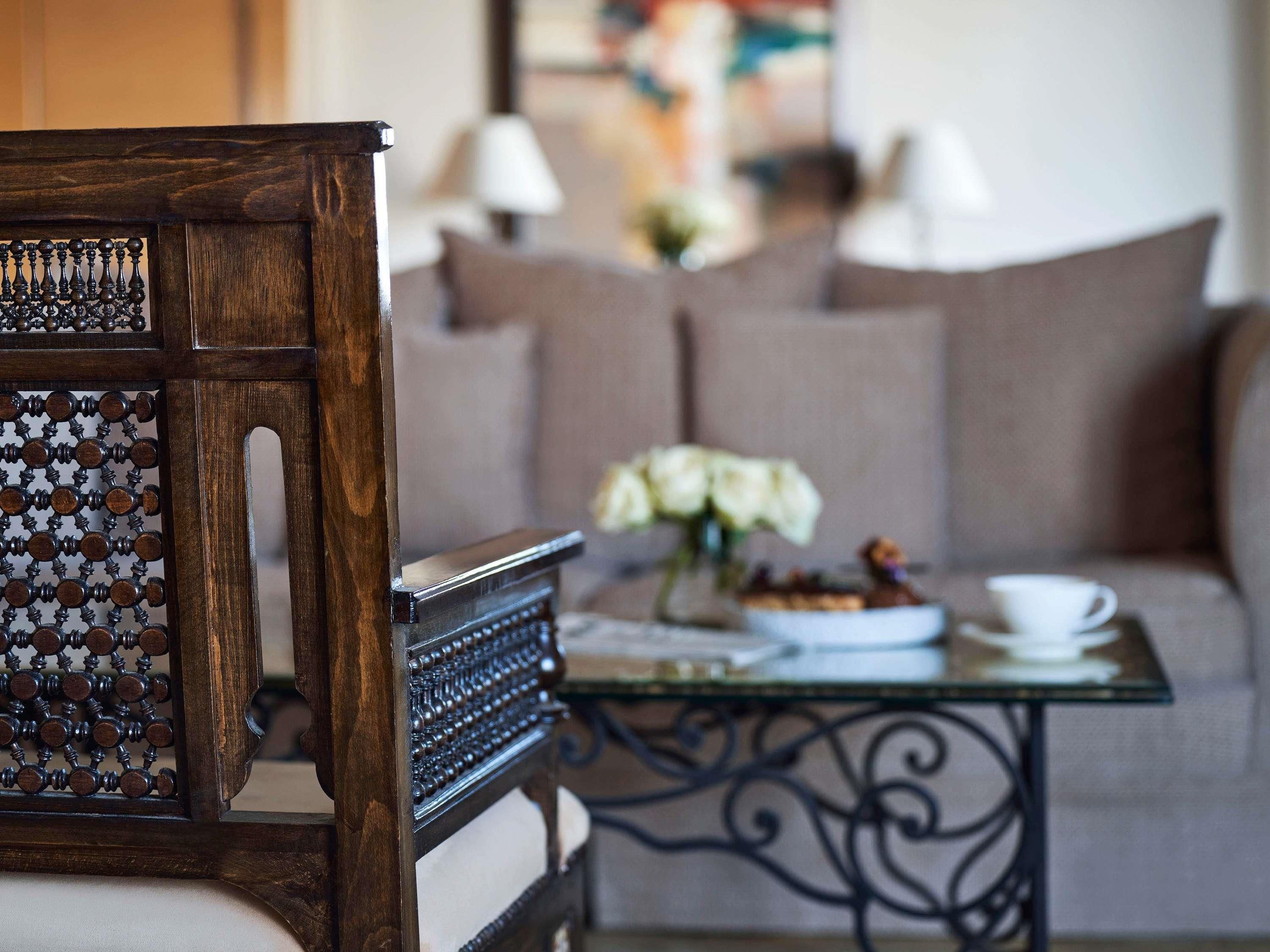 A wooden chair with intricate carvings, a glass table with a white cup and saucer, and a sofa in the background.