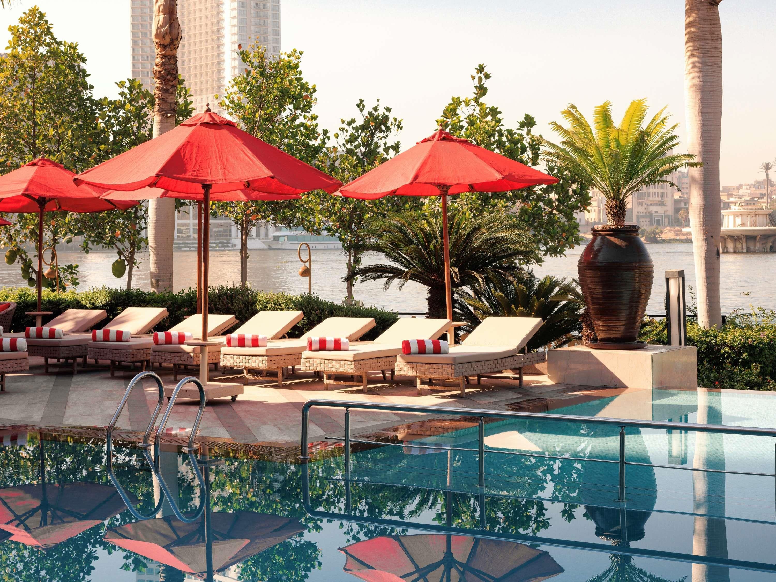 A poolside area with lounge chairs and red umbrellas, overlooking a body of water with buildings in the background.