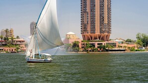 A sailboat with a white sail on a body of water, a tall building with a unique architectural design in the background, and a clear blue sky. - Sofitel Cairo Nile El Gezirah (Cairo)