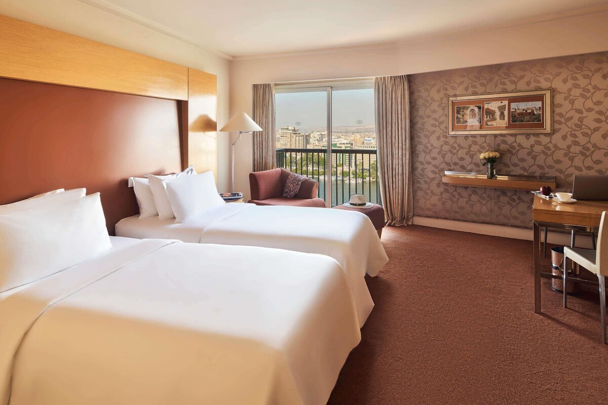 A hotel room with two beds, a desk, a chair, a lamp, a view of the city, and a patterned wall.