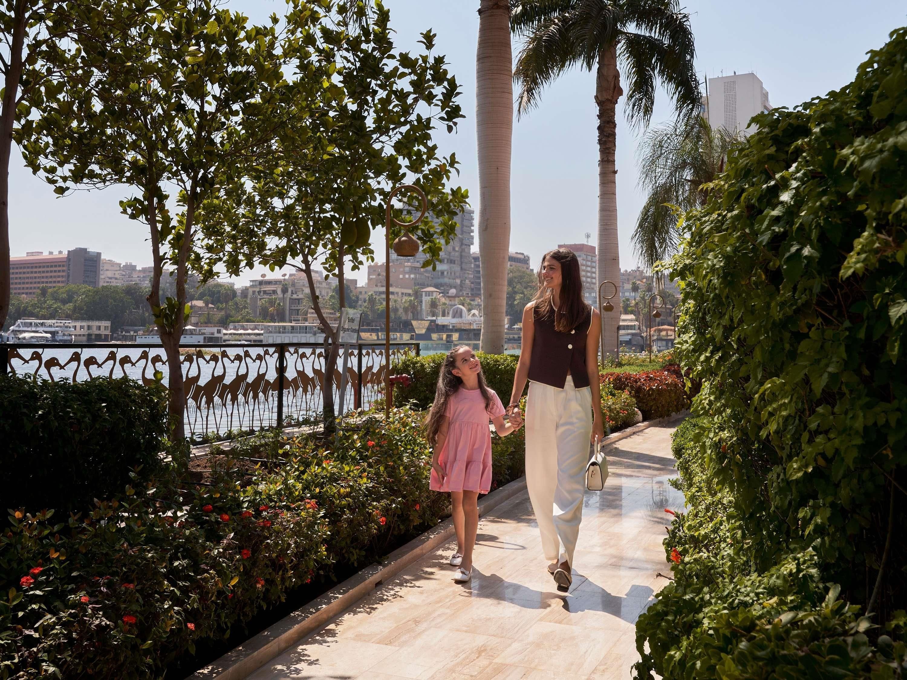 A pathway with a railing, surrounded by greenery and a view of a body of water with buildings in the background.