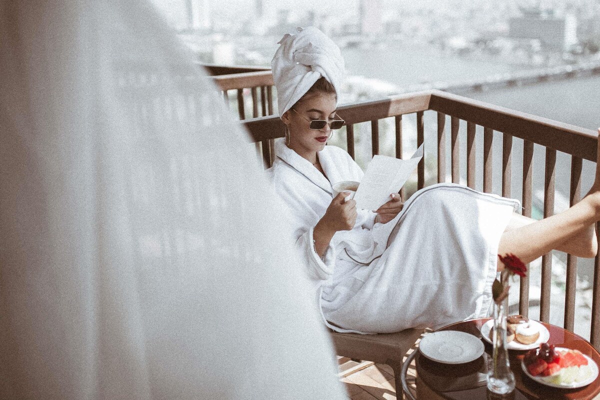 A person in a white bathrobe sitting on a wooden deck, reading a book. There is a plate with food and a glass of water on the table.