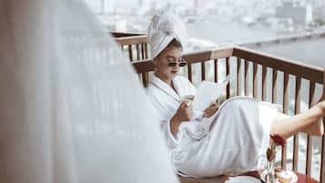 A person in a white bathrobe sitting on a wooden deck, reading a book. There is a plate with food and a glass of water on the table.