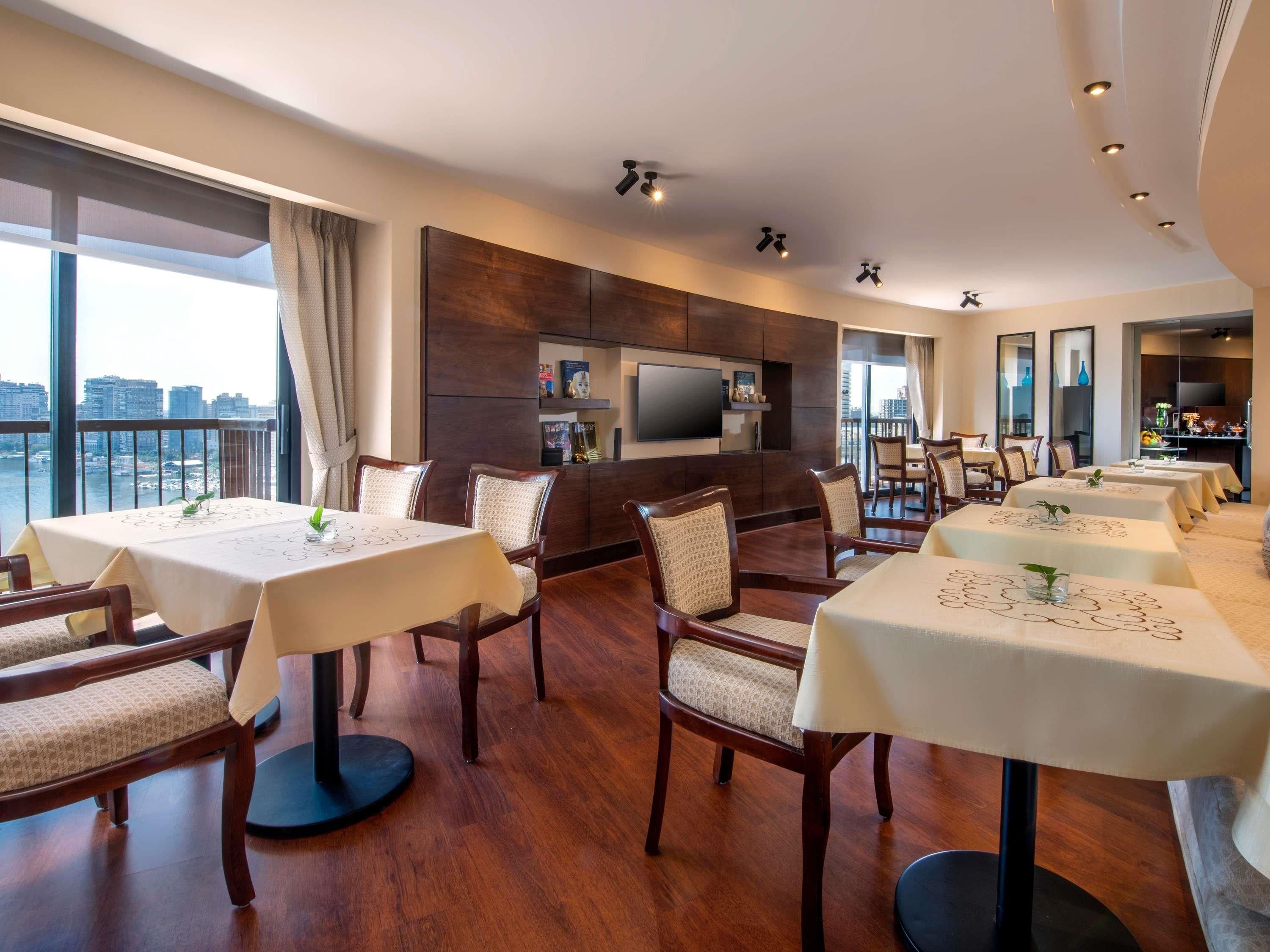 A spacious dining area with multiple tables and chairs, a flat-screen TV mounted on the wall, and a view of the cityscape through large windows.