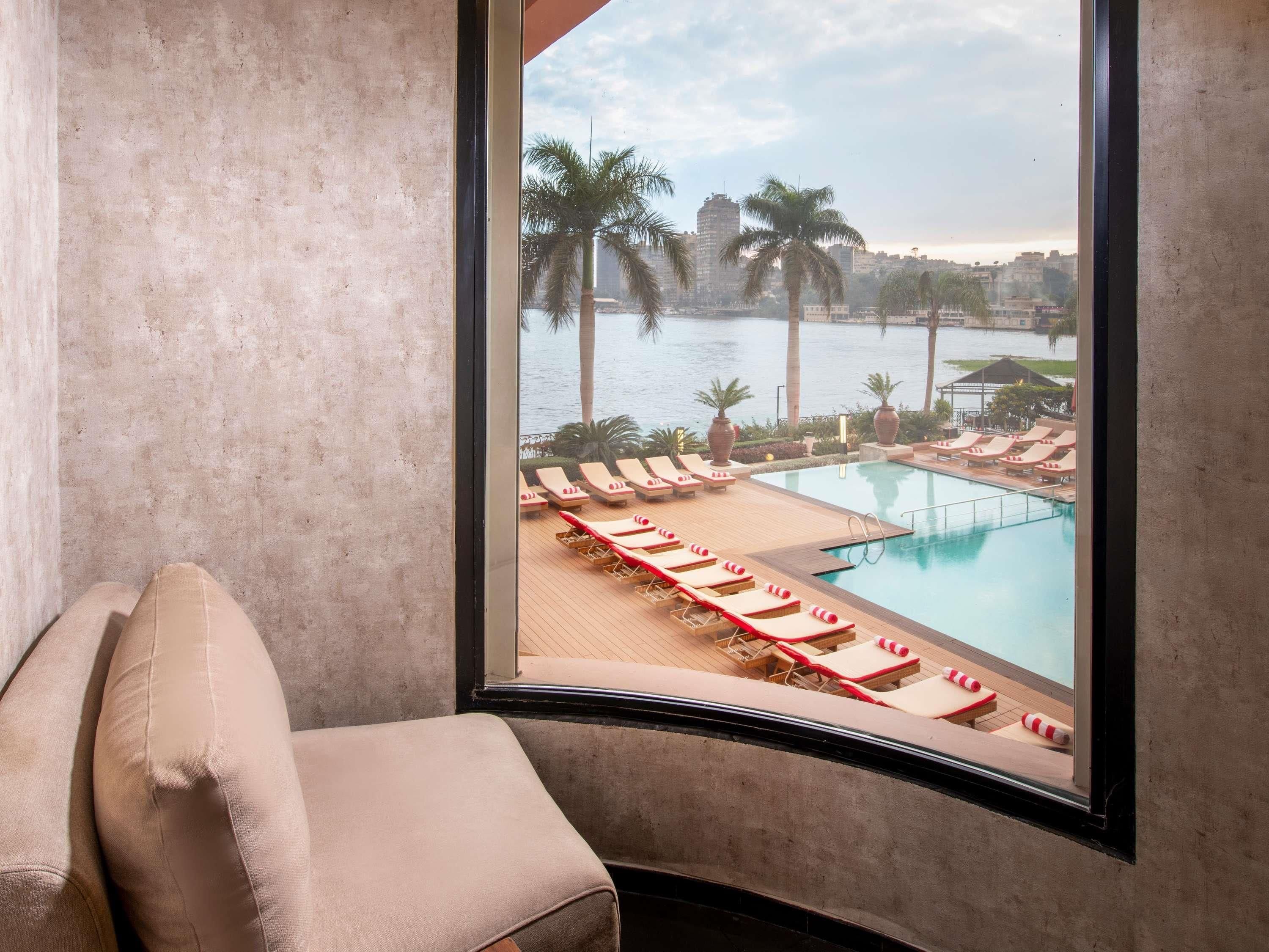 A room with a large window overlooking a pool area with sun loungers and palm trees.