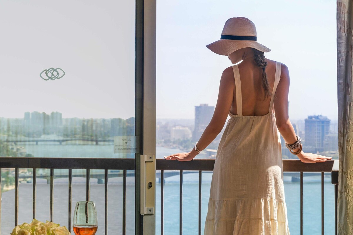 A woman in a white dress and hat standing by a balcony railing with a view of a cityscape and water.