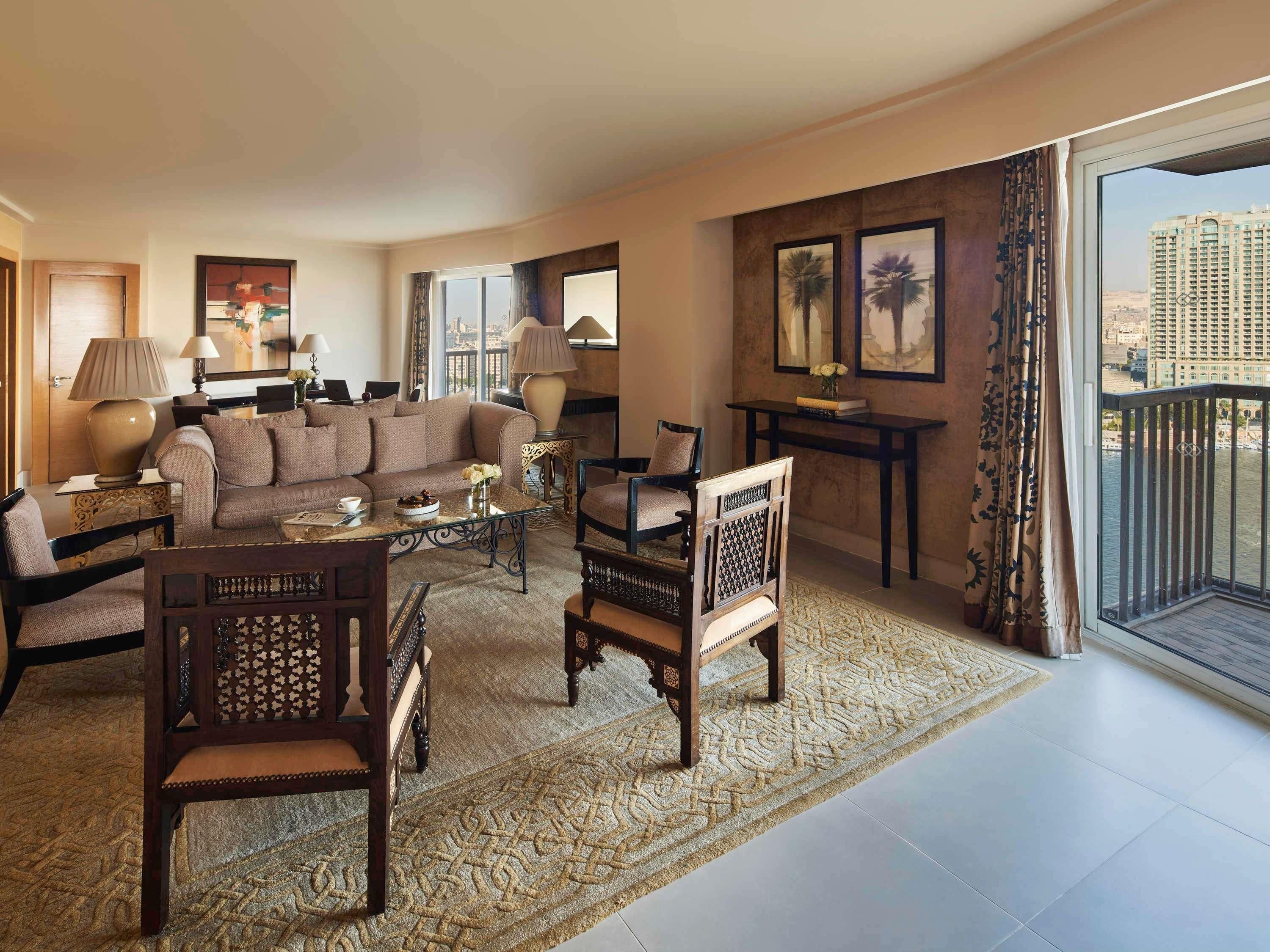 A spacious living room with a large sofa, two armchairs, a coffee table, and a small side table. There is a large window with a view of a cityscape and a balcony with a railing.