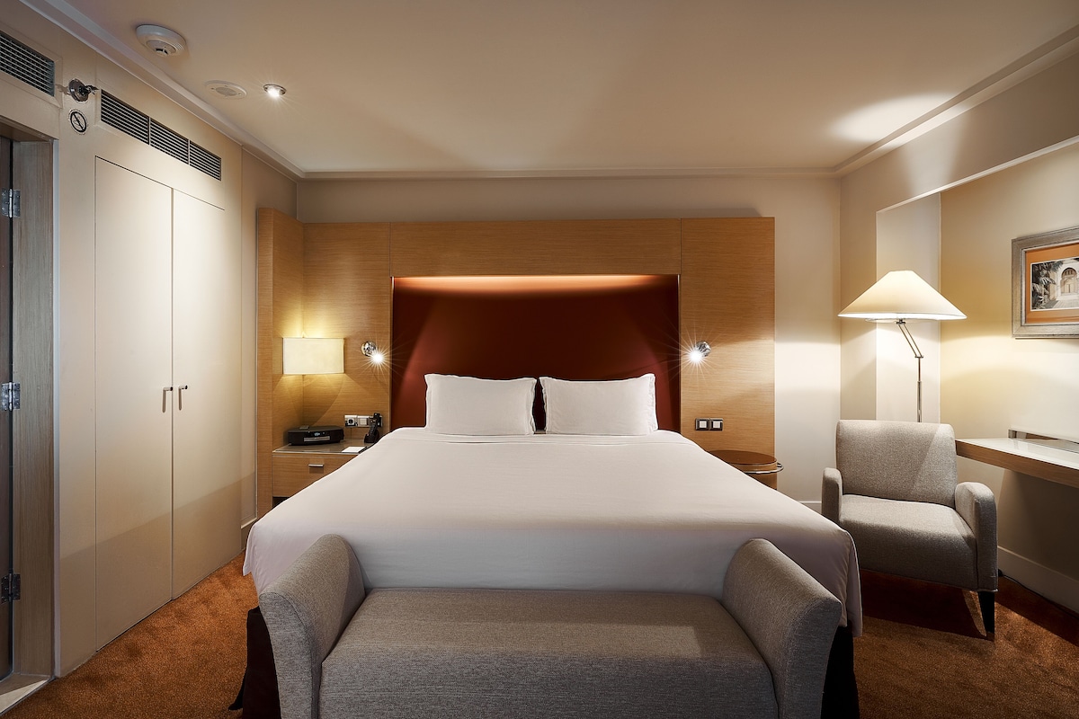 A hotel room with a large bed, a desk, a chair, and a wardrobe.
