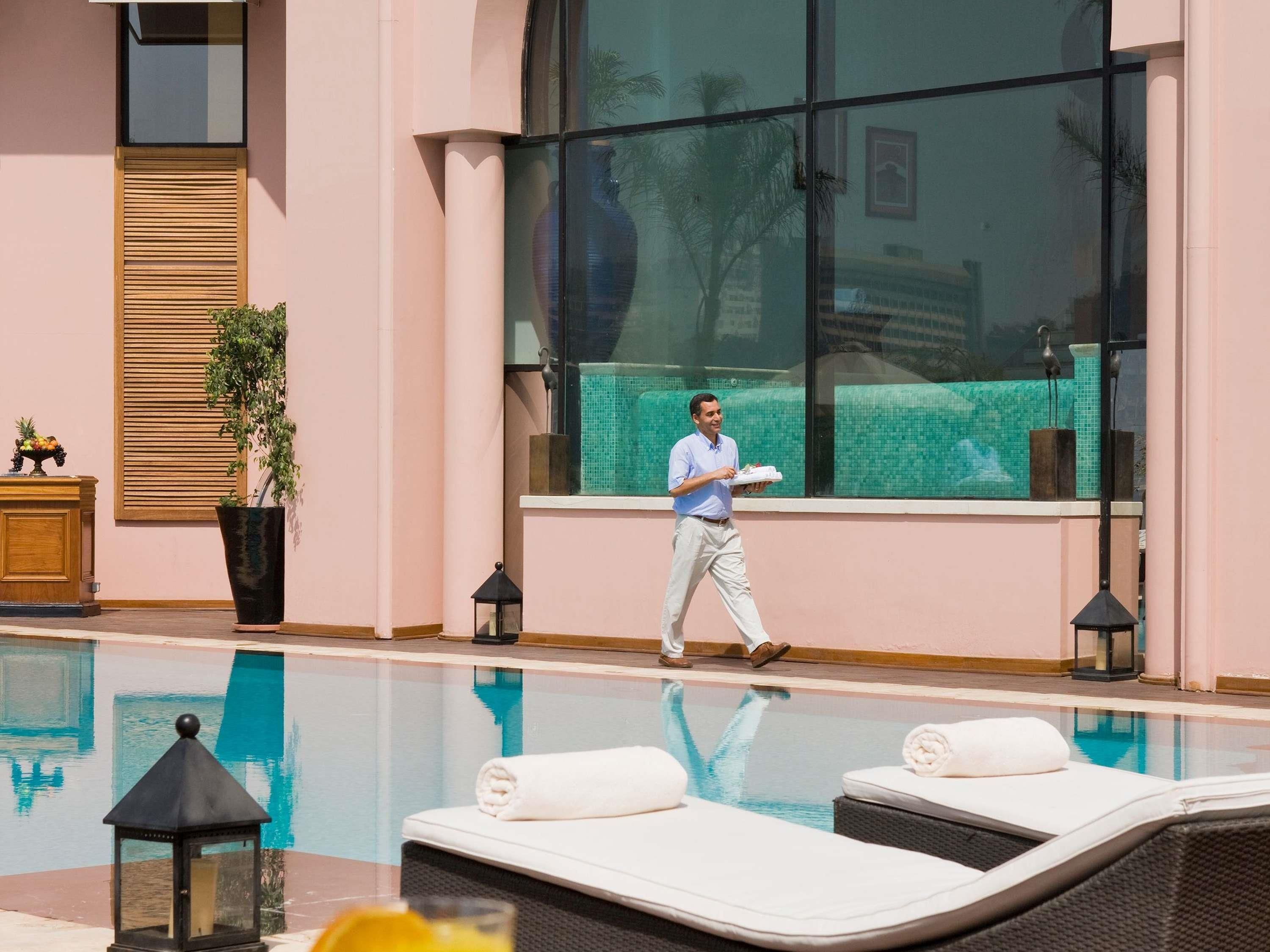 A poolside area with lounge chairs, a waiter carrying a tray, and a large window with a view of a building.