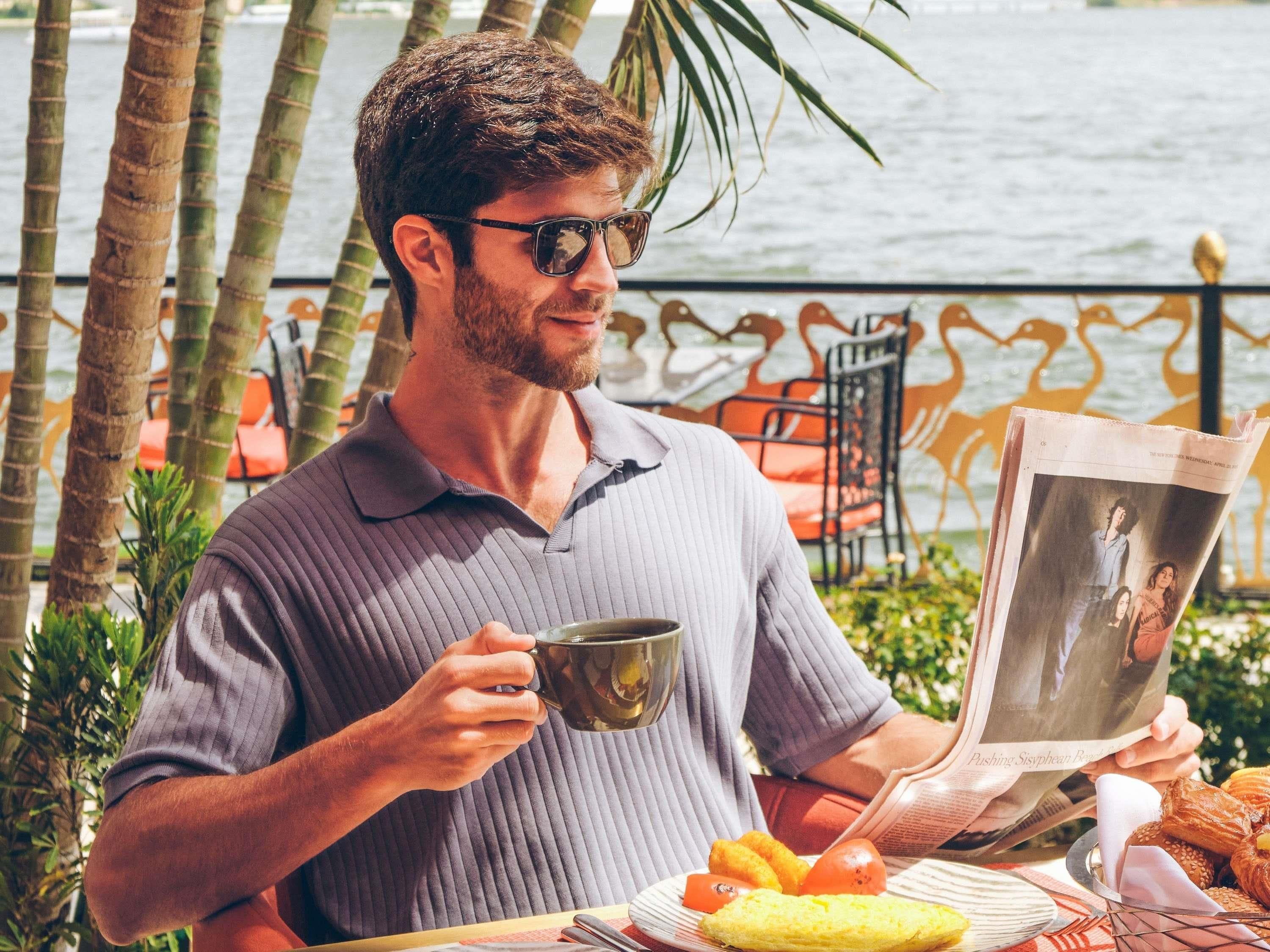A person reading a newspaper by a table with food and a drink, near a body of water and palm trees.