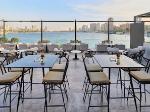 A terrace with outdoor seating, a view of the water, and city buildings in the background. - Sofitel Cairo Nile El Gezirah (Cairo)