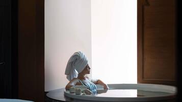 A woman in a bathtub with a towel on her head, a bottle, and a cup on the edge.