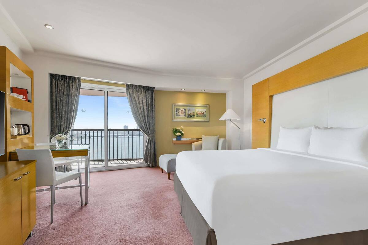 Quarto de hotel com cama, escrivaninha, cadeira e vista da cidade através de uma varanda.