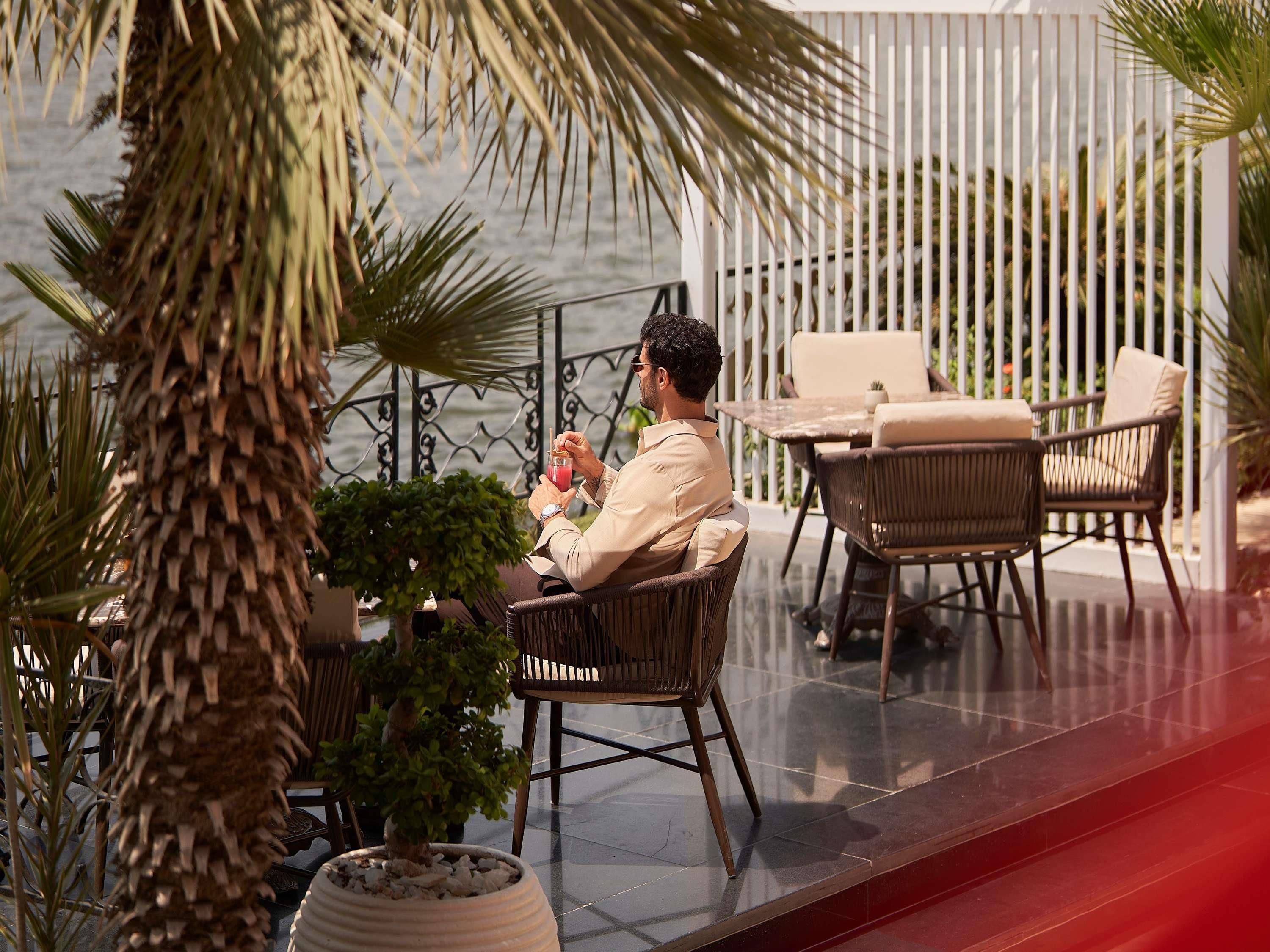 A man seated at an outdoor table with a drink, surrounded by wicker furniture and potted plants.
