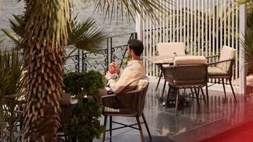 A man seated at an outdoor table with a drink, surrounded by wicker furniture and potted plants.