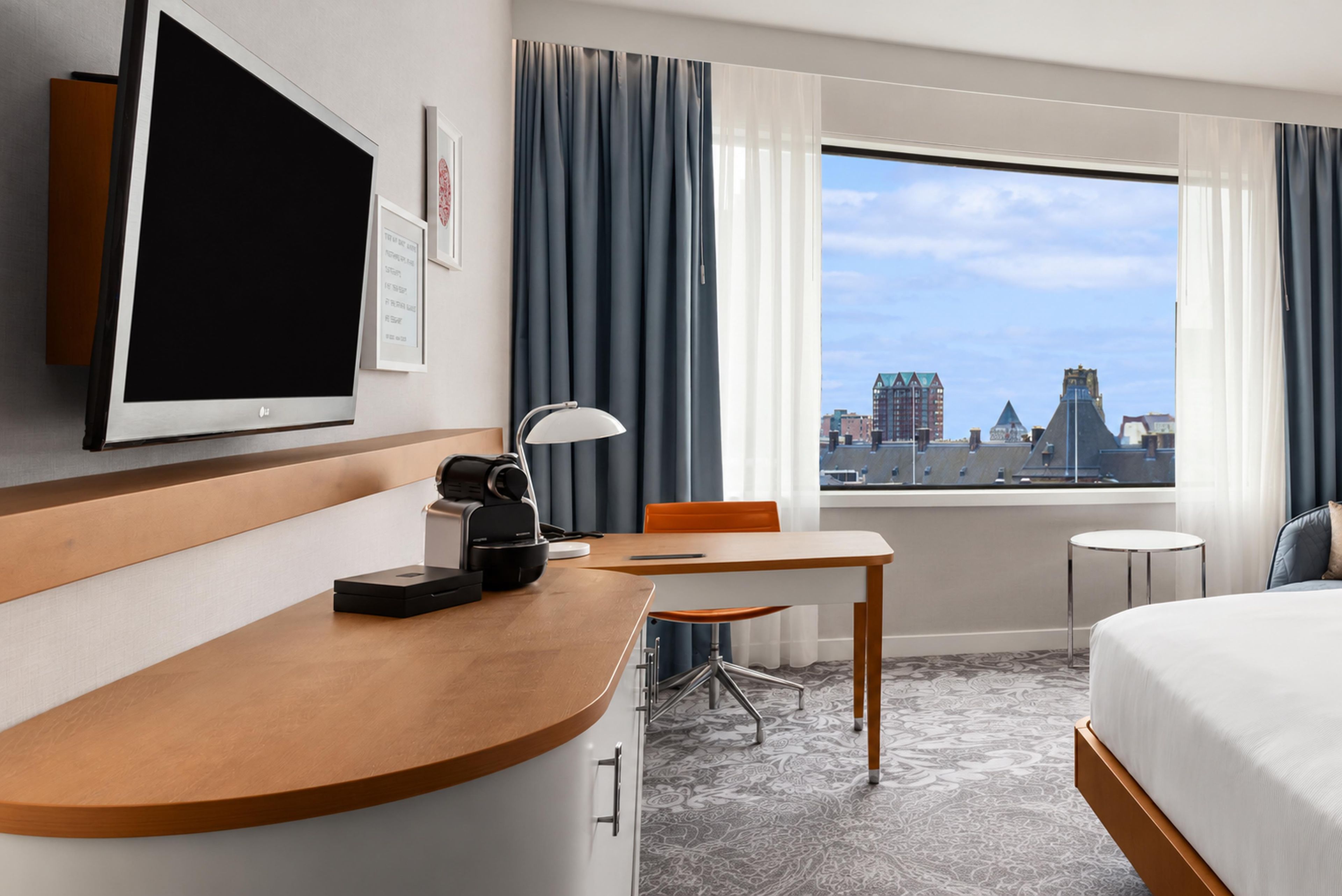 Une chambre d’hôtel avec un lit, un bureau équipé d’un ordinateur, une chaise, une télévision et une vue sur le paysage urbain.