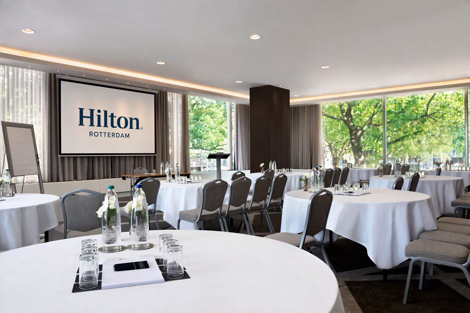 A Hilton Rotterendam conference room with multiple round tables, chairs, and a large screen displaying the hotel's name.