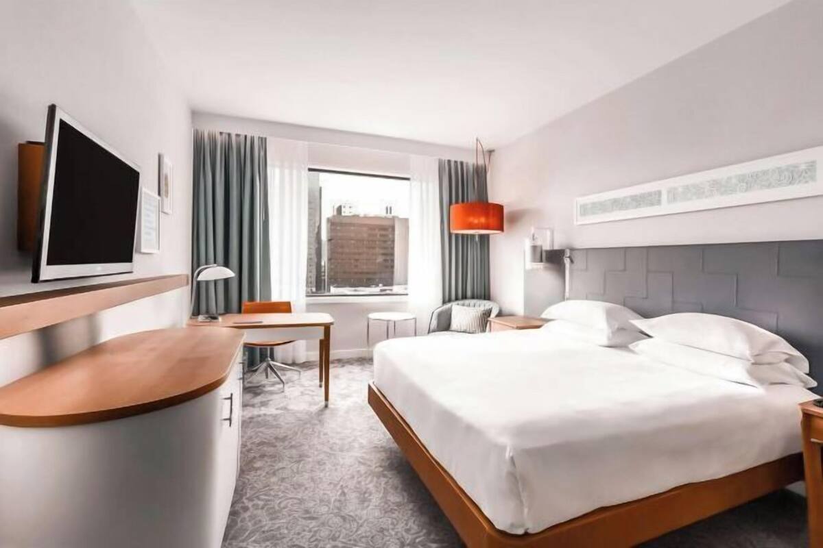 A hotel room with a bed, a desk, a chair, a TV, a window with curtains, and a city view.