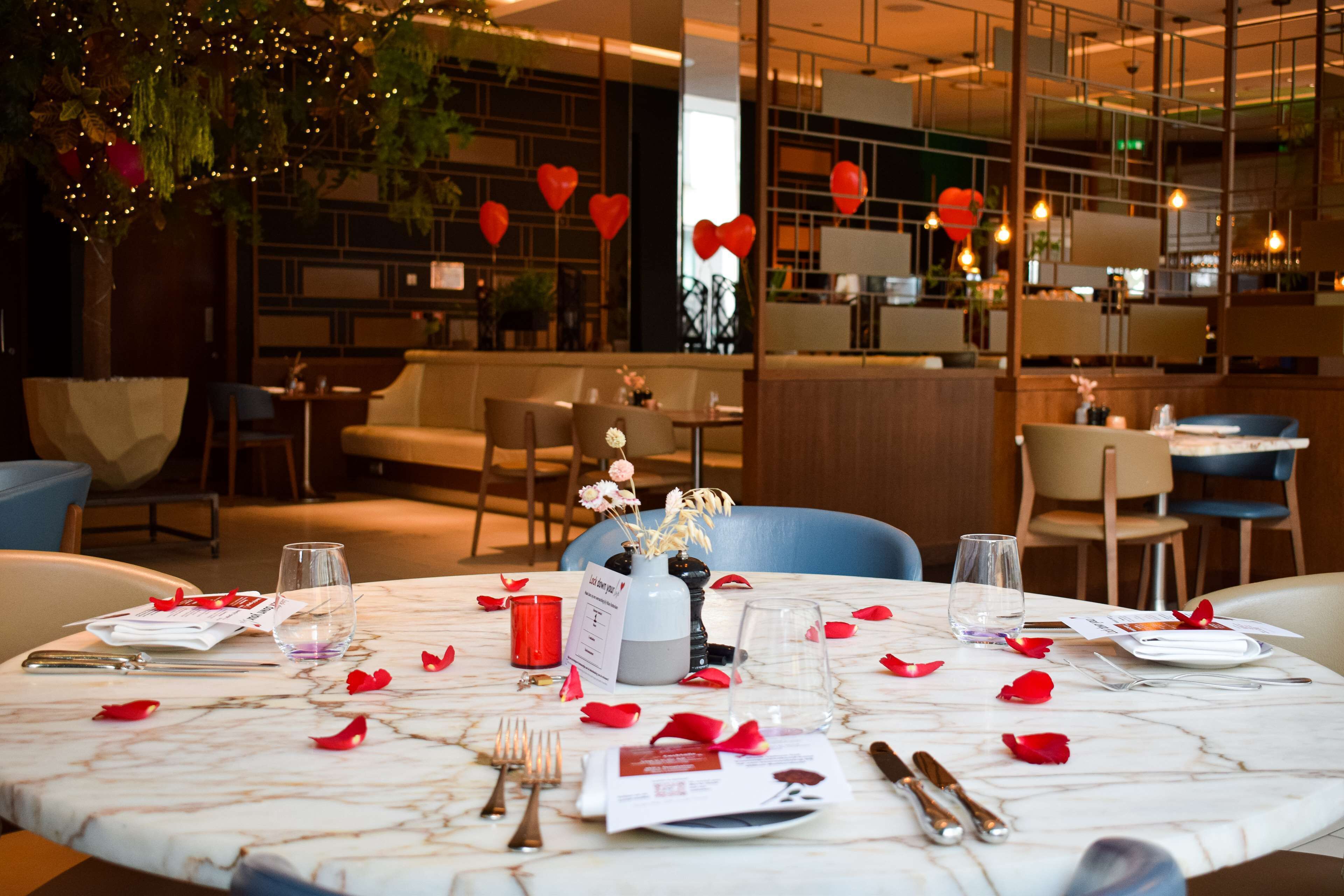 Une table dressée pour un dîner à deux, avec un menu, des verres, des couverts et un vase rempli de fleurs.