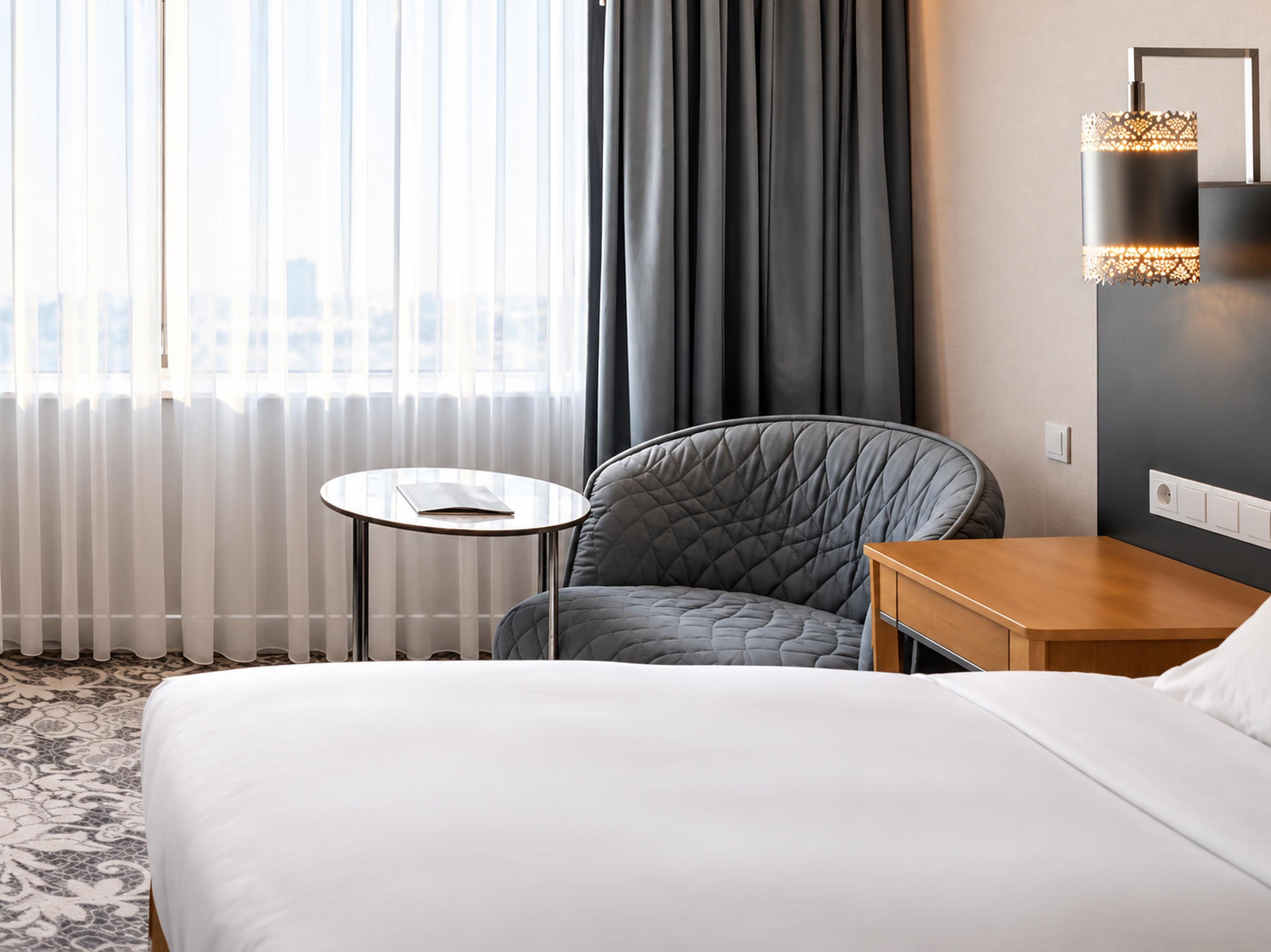 A hotel room with a bed, a round table with a book, a chair, and a wall-mounted lamp.