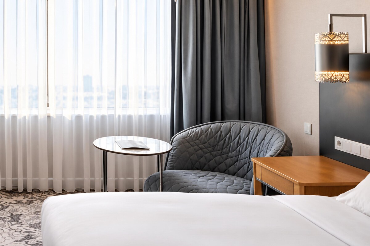 A hotel room with a bed, a round table with a book, a chair, and a wall-mounted lamp.