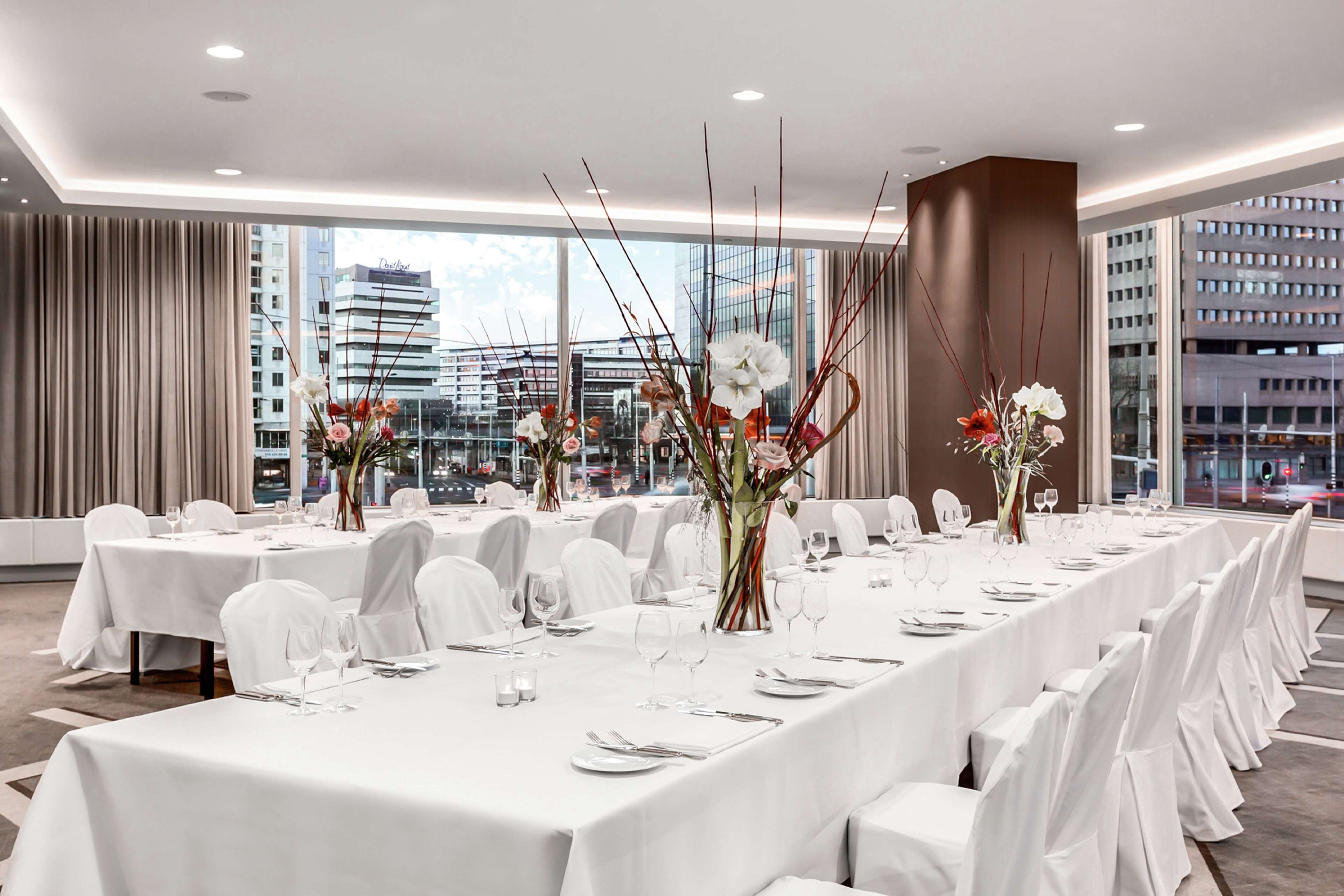 A hotel dining area with long tables set for a formal event, large windows offering a city view, and modern interior design.