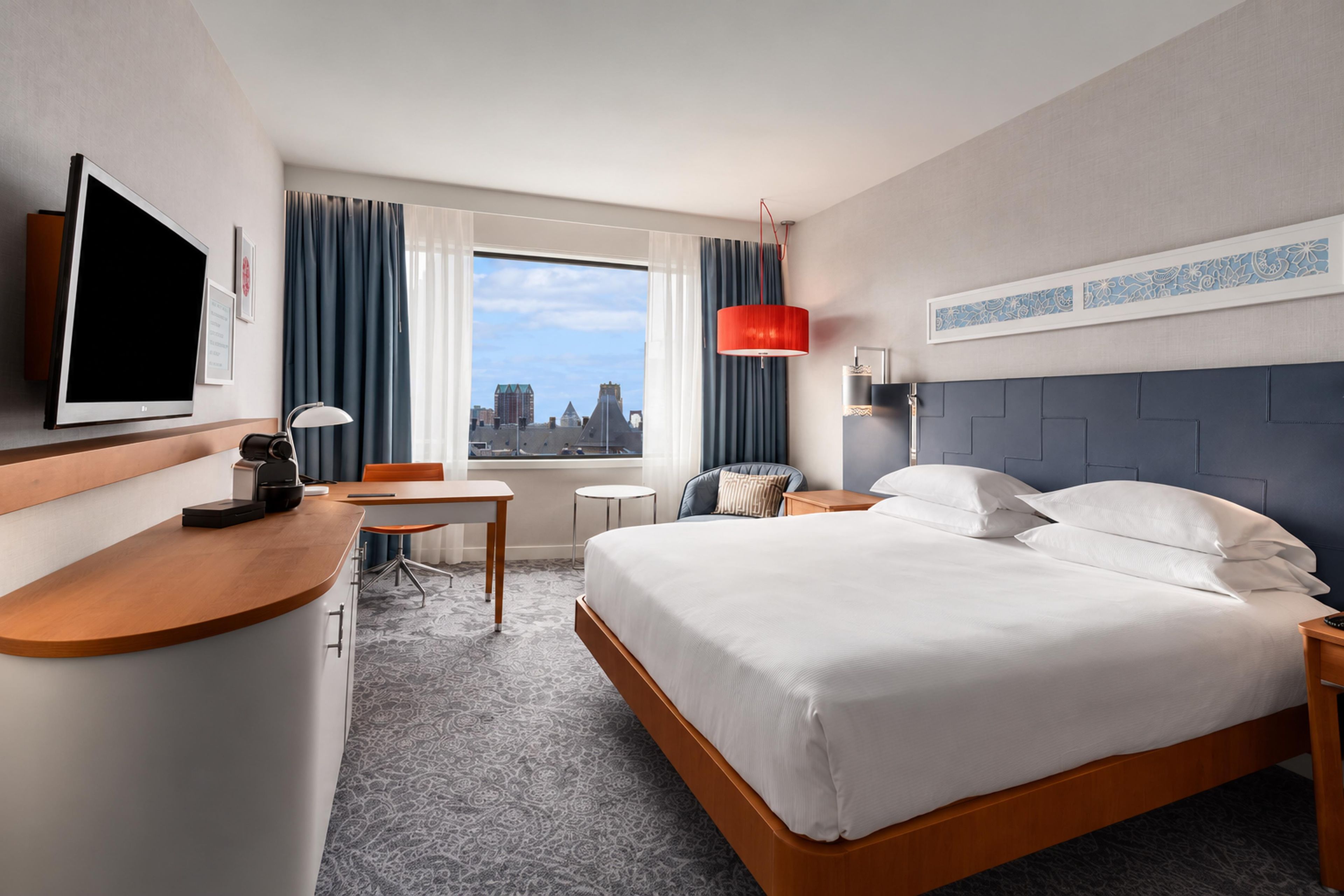 A hotel room with a bed, a desk, a chair, a TV, a window with curtains, and a city view.
