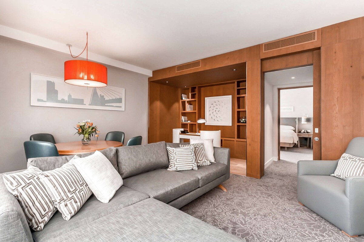 Een moderne woonkamer met een grijze hoekbank, een houten inbouwkast en een rode hanglamp.