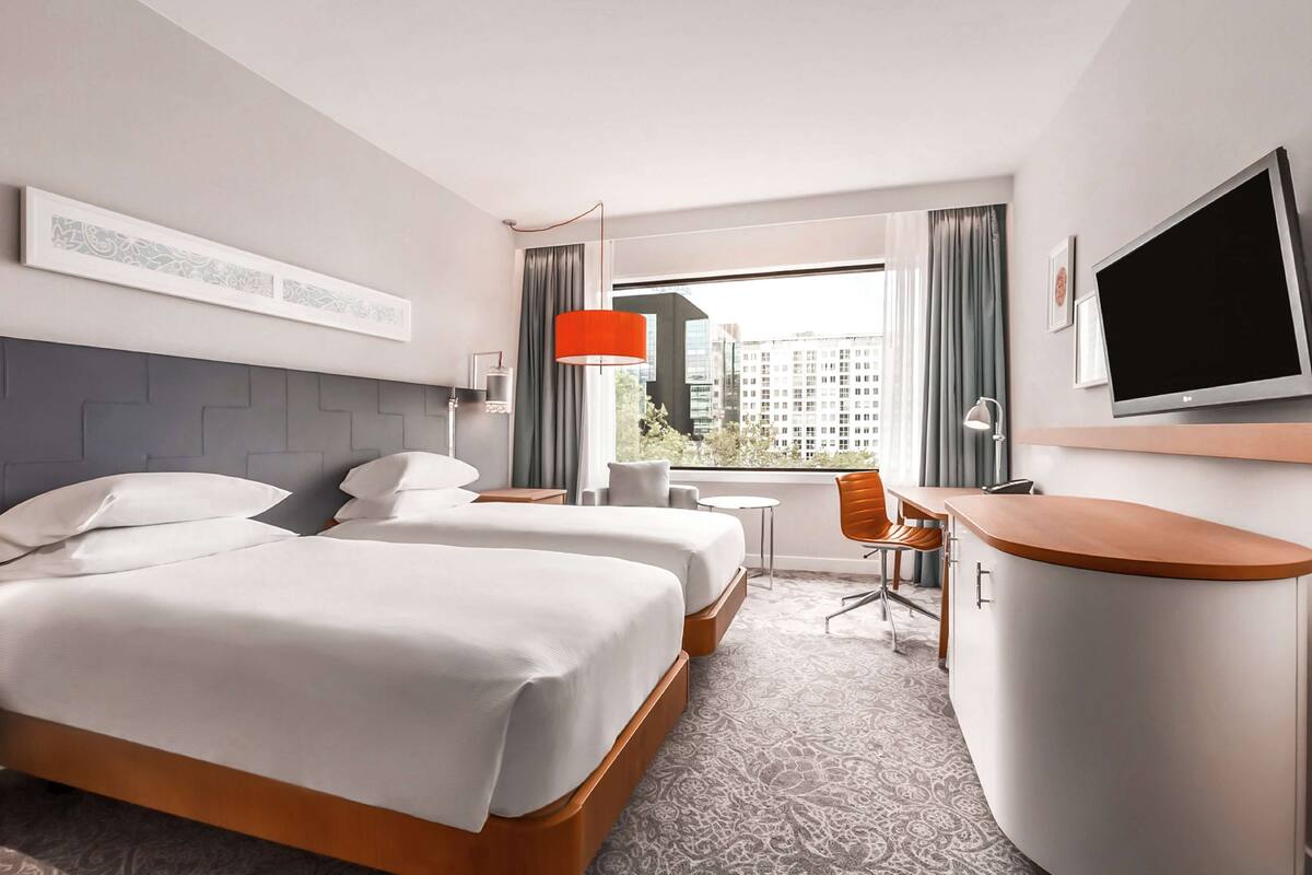 A hotel room with two beds, a desk, a chair, a TV, and a window with a city view.